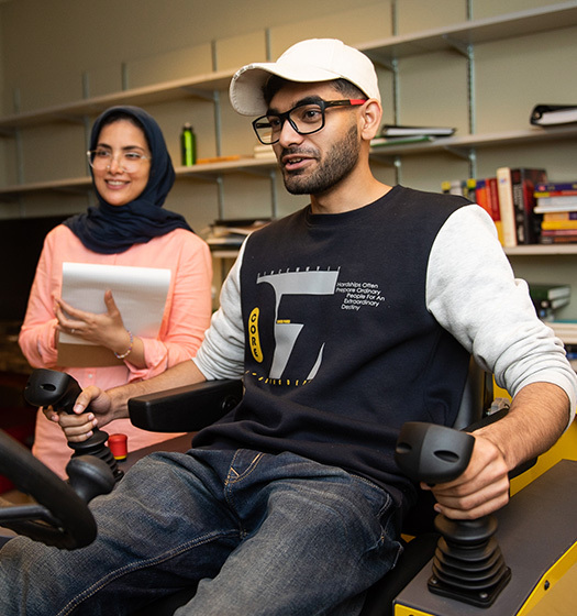 A man sitting on a gaming simulator with a woman next to him taking notes on a writing pad