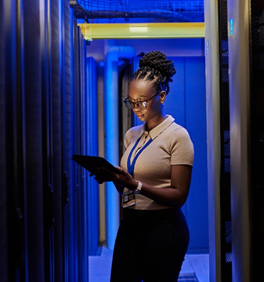 A woman standing between server racks in a data center, holding a tablet and wearing a lanyard badge. 