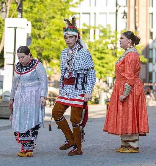 Three people dressed in traditional Native American regalia standing outdoors on a paved area with greenery and buildings in the background.