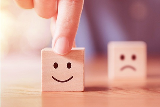 A hand holds up a wooden tile with a smiley face printed on it on a table. In the background, out of focus, is a tile with a sad face.